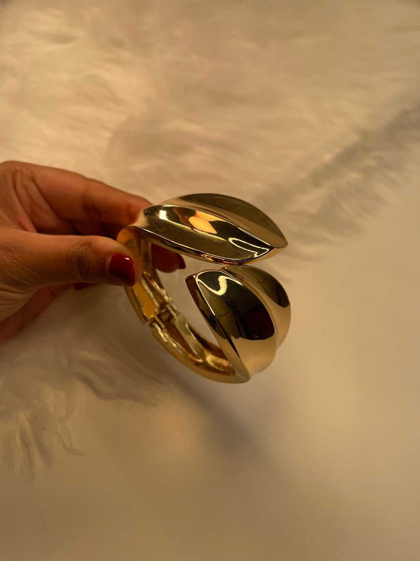 Gold bracelet held by a hand with red nail polish against a neutral background