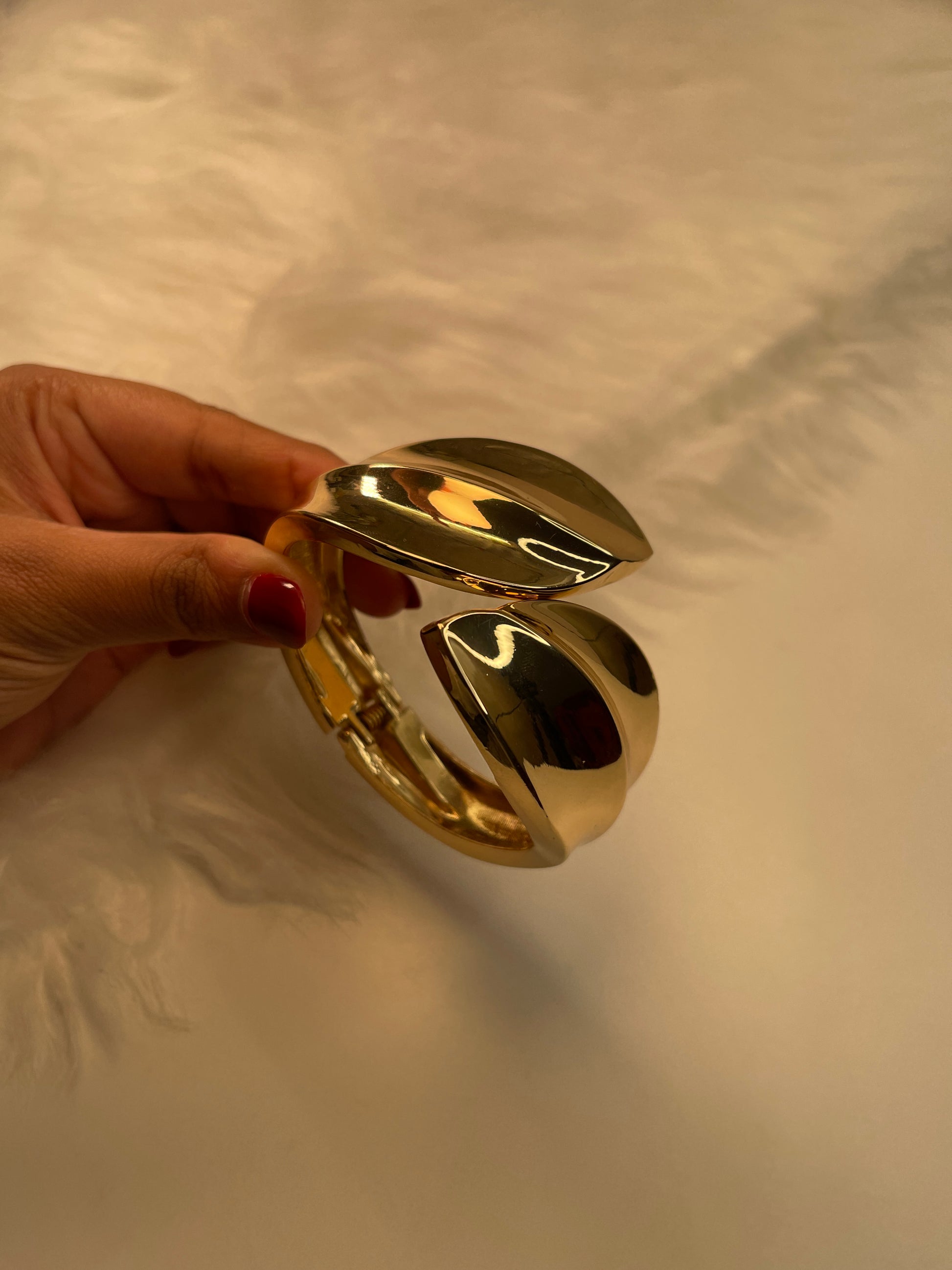 Gold bracelet held by a hand with red nail polish against a neutral background