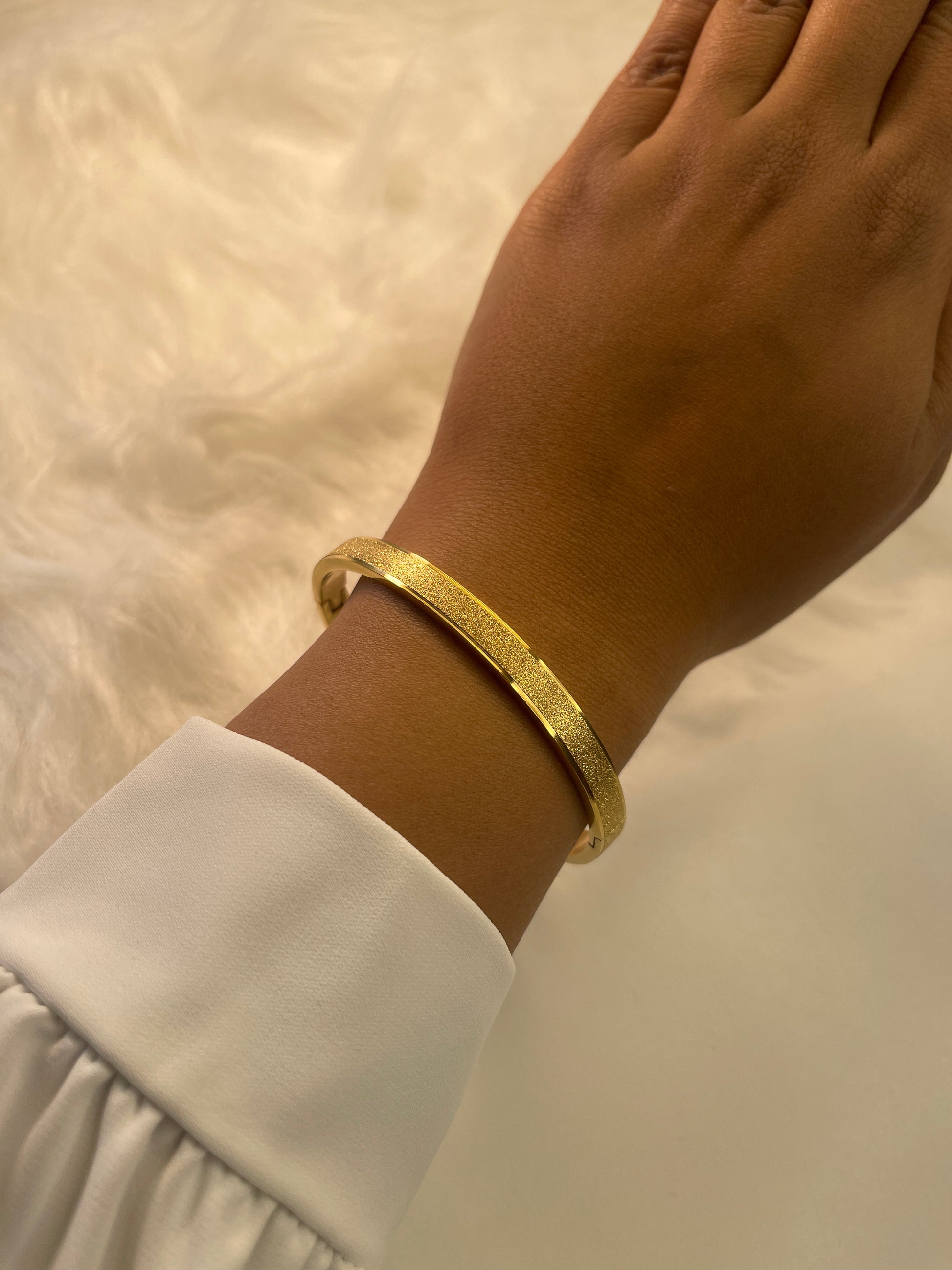 Gold bracelet on a wrist with a white sleeve against a neutral background