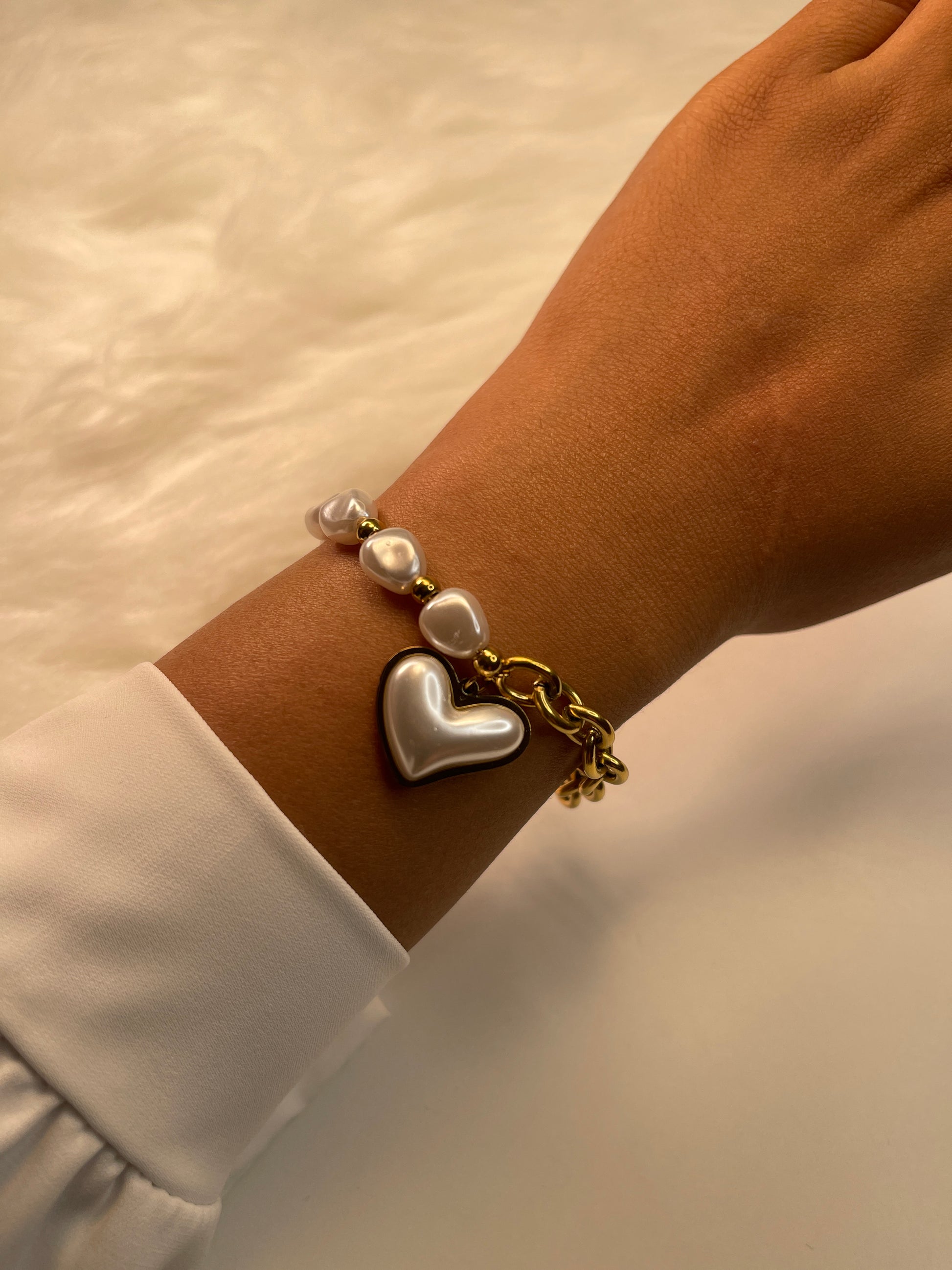 Gold bracelet with heart and pearl charms on a wrist against a neutral background