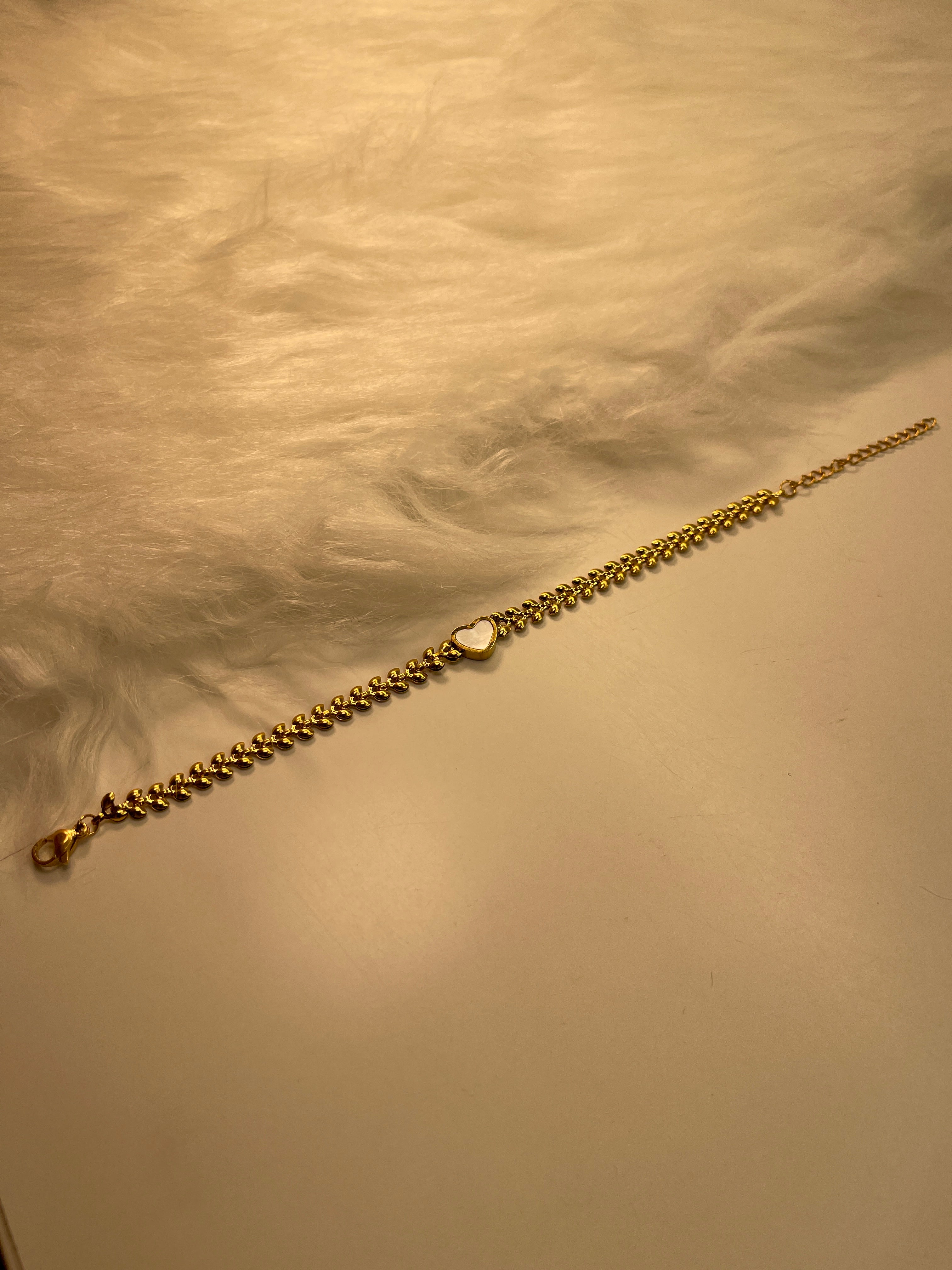 Gold chain bracelet with a white heart pendant on a textured beige surface